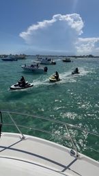 View from a boat bow over clear turquoise coastal water with several boats and three jet skis speeding past under a towering white cloud on a sunny day.