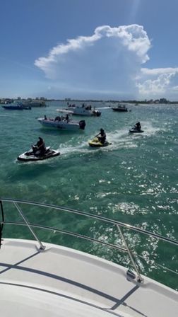 View from a boat bow over clear turquoise coastal water with several boats and three jet skis speeding past under a towering white cloud on a sunny day.