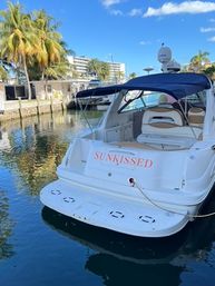 White motor yacht named 'SUNKISSED' moored at a sunny tropical marina dock with palm trees, waterfront buildings, calm reflective water and blue sky.