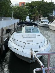 White motor yacht docked at a marina, bow facing the camera, flanked by neighboring boats with waterfront trees and apartment buildings in the background.