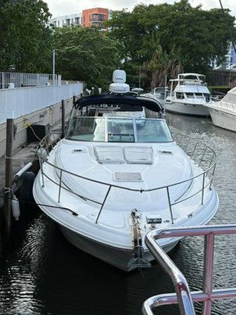 White motor yacht docked at a marina, bow facing the camera, flanked by neighboring boats with waterfront trees and apartment buildings in the background.