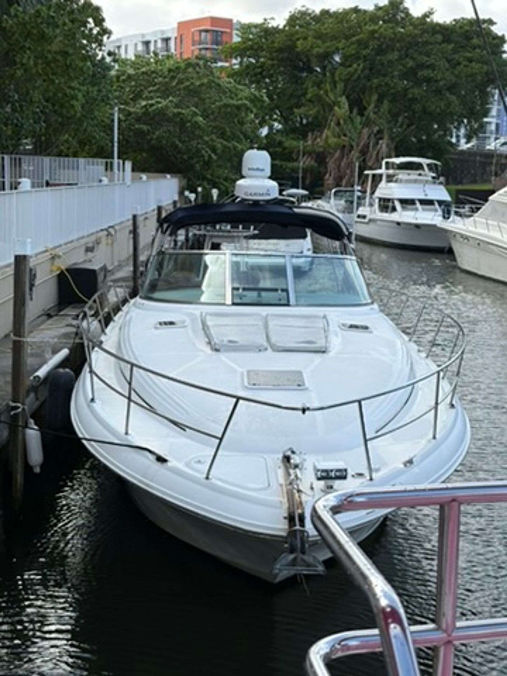 White motor yacht docked at a marina, bow facing the camera, flanked by neighboring boats with waterfront trees and apartment buildings in the background.