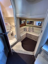 Cozy compact yacht cabin interior with cream curved walls, L-shaped V-berth lounge and striped throw pillows, mirrored bulkhead, carpeted floor and adjacent small marine head with open toilet.