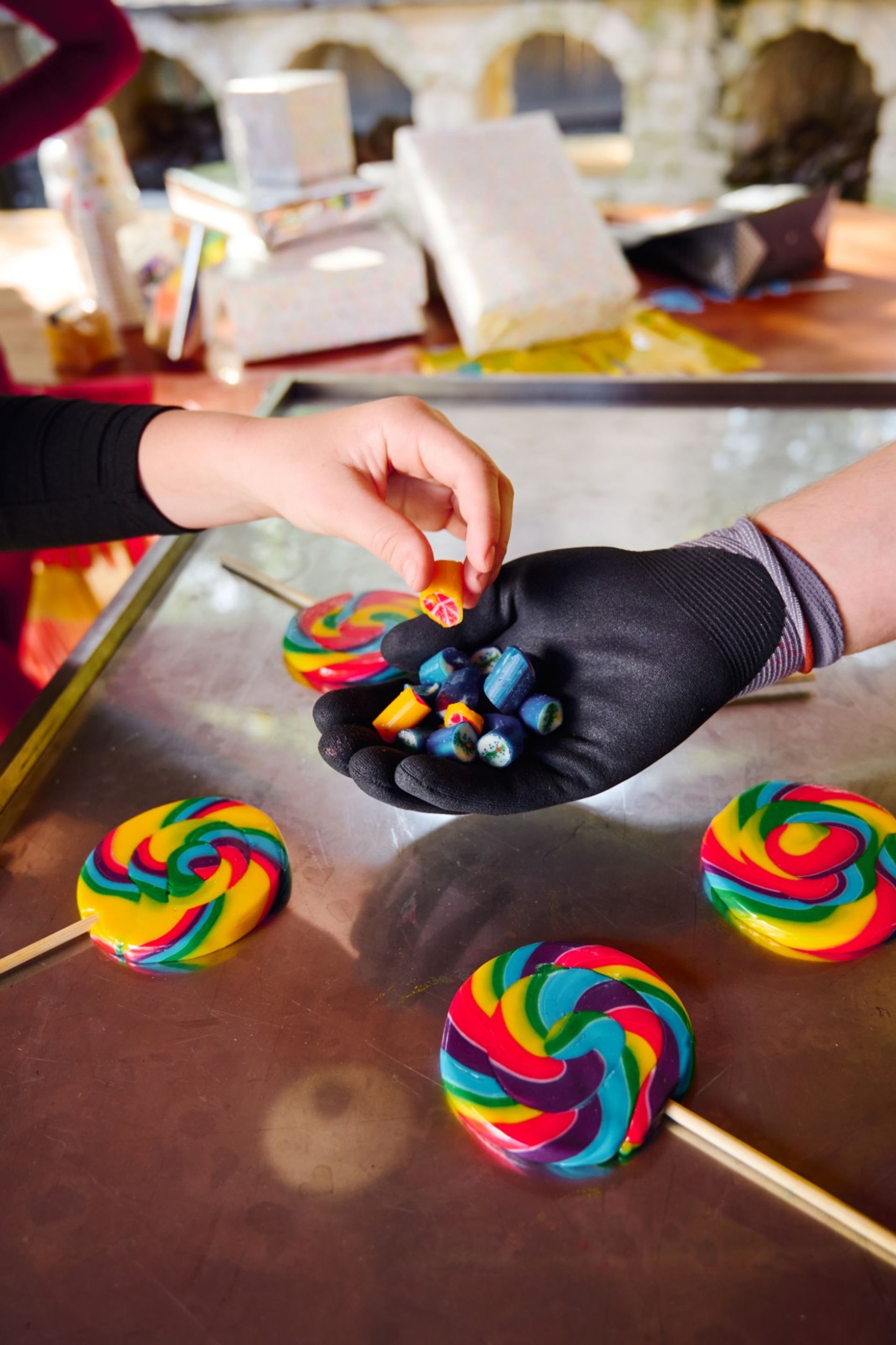 Gloved vendor hand holding colorful handmade hard candy pieces as another hand picks one, with bright rainbow swirl lollipops on a metal counter.
