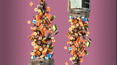 Two glass jars tipping and spilling colorful striped and fruit-patterned rock candies frozen mid-air against a pink background