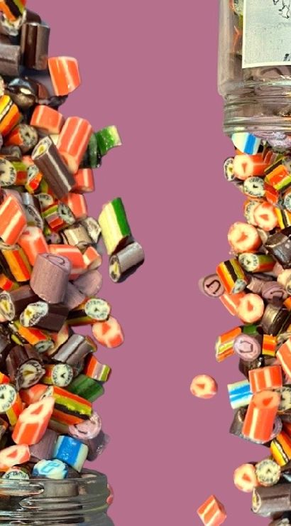 Two glass jars tipping and spilling colorful striped and fruit-patterned rock candies frozen mid-air against a pink background