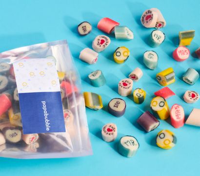 Bright turquoise background with scattered colorful rock-candy slices printed with hearts, 'MR'/'MRS' and "Just Married", and a clear resealable bag at left
