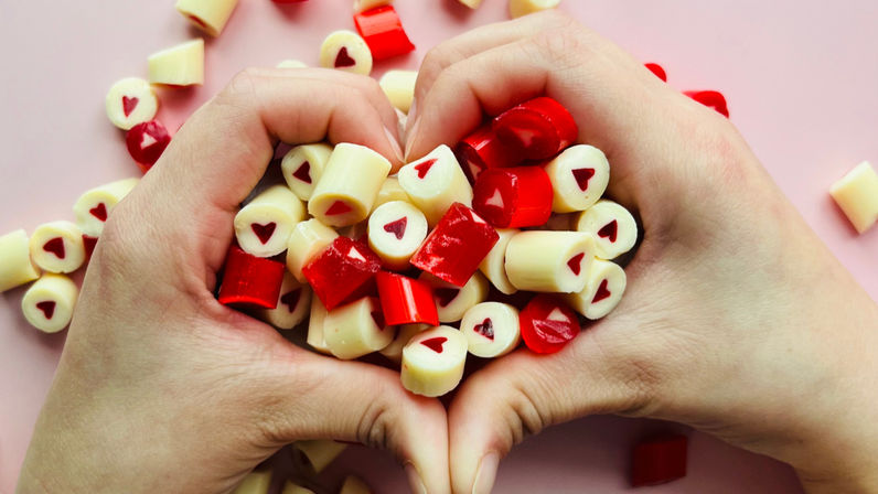 Hands forming a heart holding red-and-white hard candies with tiny red heart centers on a pink background, Valentine candy close-up