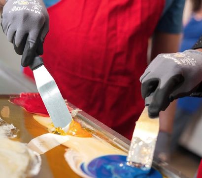 Close-up of gloved hands in a red apron using metal spatulas to swirl bright blue, yellow and red pulled sugar candy on a stainless-steel table in an artisanal candy shop.
