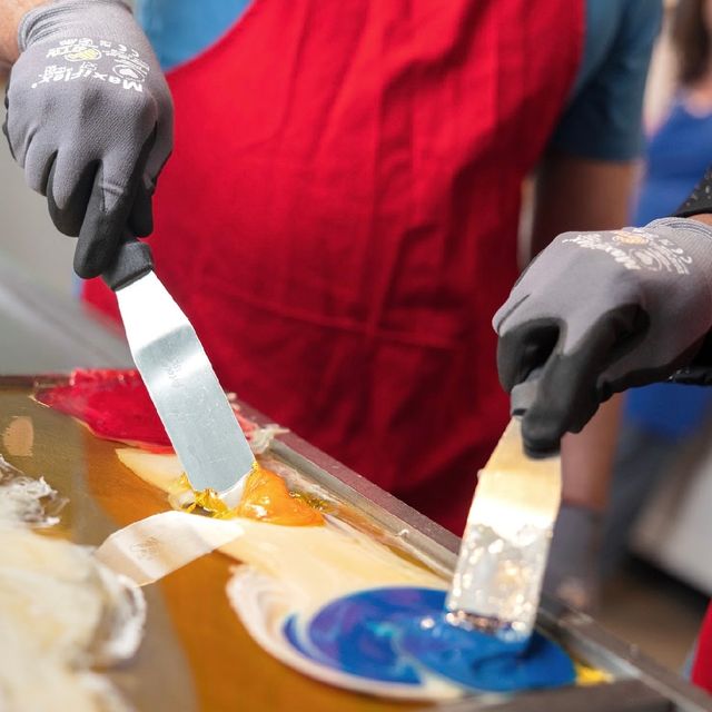 Close-up of gloved hands in a red apron using metal spatulas to swirl bright blue, yellow and red pulled sugar candy on a stainless-steel table in an artisanal candy shop.