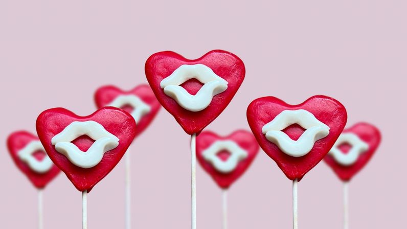 Red heart-shaped lollipops with white candy lips on sticks against a soft pink background, playful Valentine's Day sweets