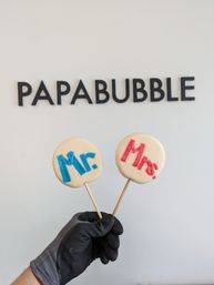 Gloved hand holding two round white candy lollipops on wooden sticks labeled “Mr.” in blue and “Mrs.” in red, photographed against a white confectionery wall with black lettering.
