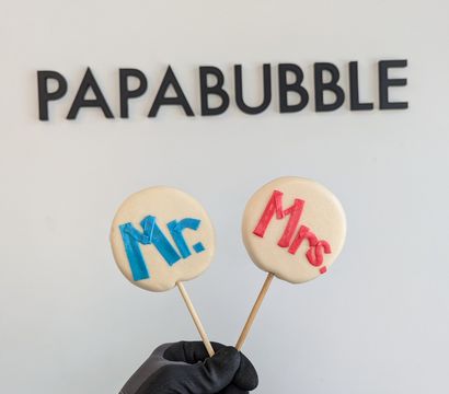 Gloved hand holding two round white candy lollipops on wooden sticks labeled “Mr.” in blue and “Mrs.” in red, photographed against a white confectionery wall with black lettering.