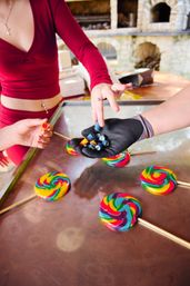 Close-up of hands sharing bright mini candies and rainbow swirl lollipops on a metal counter at an outdoor market candy stall.