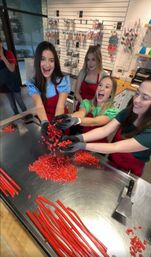 Four smiling women in aprons tossing chopped red candy over a stainless-steel worktable with long red candy rods and a spatula in an artisan candy-making workshop