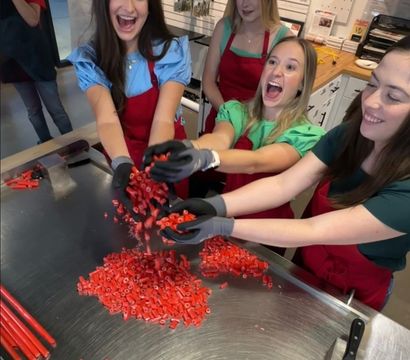 Four smiling women in aprons tossing chopped red candy over a stainless-steel worktable with long red candy rods and a spatula in an artisan candy-making workshop