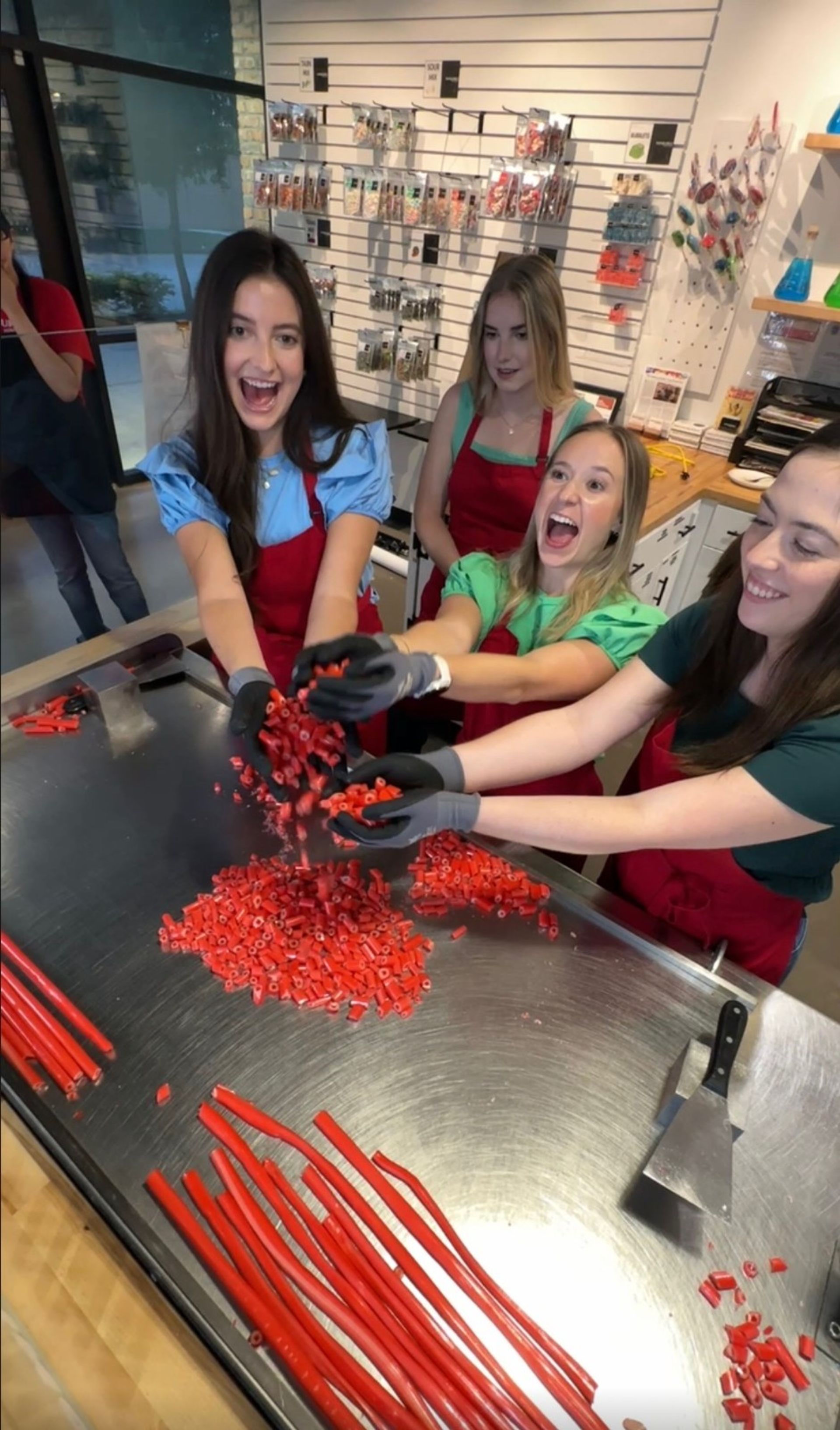 Four smiling women in aprons tossing chopped red candy over a stainless-steel worktable with long red candy rods and a spatula in an artisan candy-making workshop