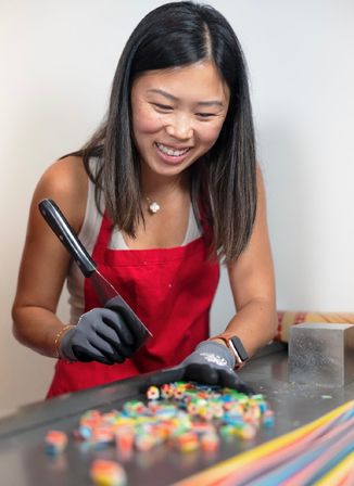 Smiling woman in a red apron and gloves slicing colorful handmade hard candy with a large knife on a worktable in a candy workshop.