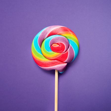 Bright rainbow swirl lollipop on a purple background, close-up flat-lay of colorful candy on a wooden stick.
