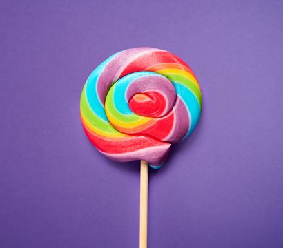 Bright rainbow swirl lollipop on a purple background, close-up flat-lay of colorful candy on a wooden stick.