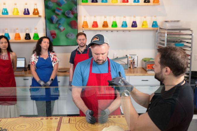Candy-making workshop scene with students in red and blue aprons watching a man shape candy at a glass counter while an instructor demonstrates, colorful conical flasks displayed on shelves in the background.