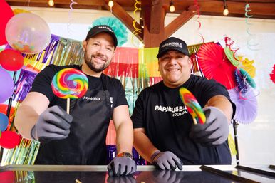 Two smiling people in black shirts and gloves hold rainbow swirled lollipops toward the camera against a colorful party backdrop of balloons, paper fans and hanging streamers.