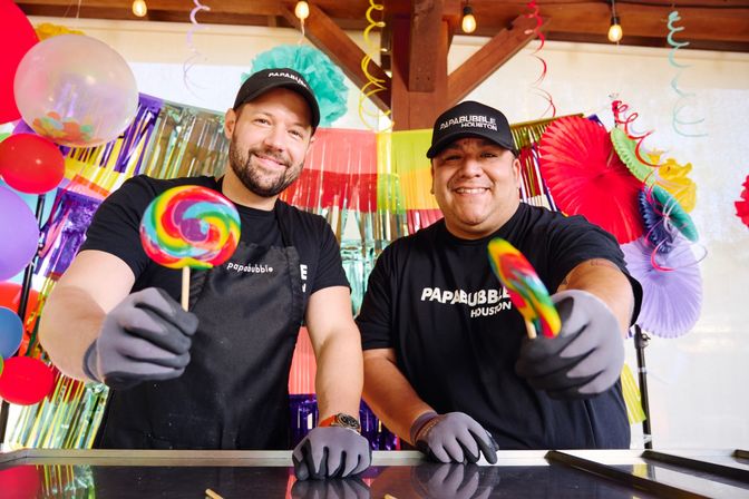 Two smiling people in black shirts and gloves hold rainbow swirled lollipops toward the camera against a colorful party backdrop of balloons, paper fans and hanging streamers.