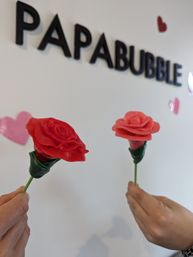 Two hands holding red and coral rose-shaped candy lollipops in a candy shop interior, blurred black wall signage and pink heart decals on a white background.
