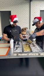 Two vendors wearing Santa hats and gloves making rolled ice cream on a chilled metal slab, pouring a yellow mixture and working with spatulas at a festive outdoor food stall against a white brick wall.