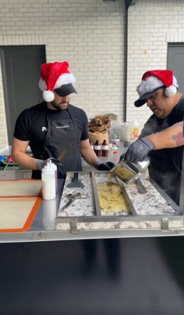 Two vendors wearing Santa hats and gloves making rolled ice cream on a chilled metal slab, pouring a yellow mixture and working with spatulas at a festive outdoor food stall against a white brick wall.
