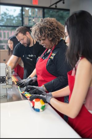 People in red aprons and gloves shaping colorful striped candy on a stainless counter during an artisan candy-making workshop in a candy shop