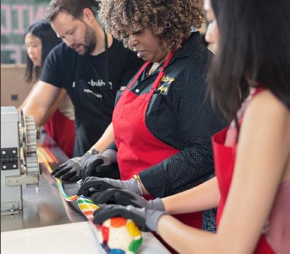 People in red aprons and gloves shaping colorful striped candy on a stainless counter during an artisan candy-making workshop in a candy shop