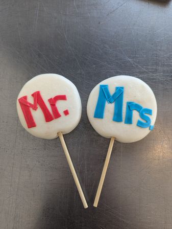 Two round white cookie pops on wooden sticks decorated with red "Mr." and blue "Mrs." fondant letters, styled as wedding favors on a scratched stainless-steel countertop