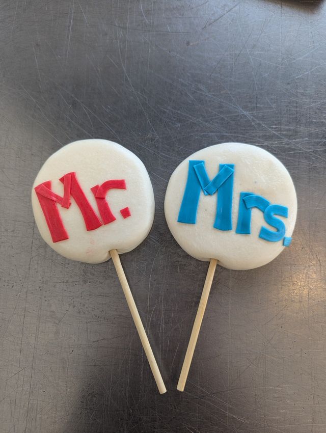 Two round white cookie pops on wooden sticks decorated with red "Mr." and blue "Mrs." fondant letters, styled as wedding favors on a scratched stainless-steel countertop
