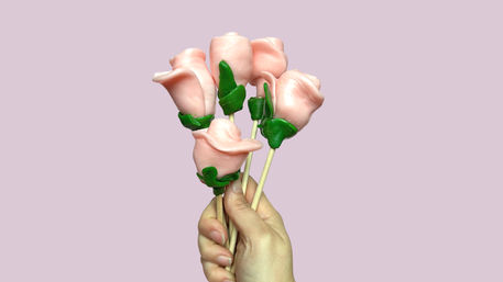 Close-up of a hand holding a bouquet of five pink candy rose lollipops with green fondant leaves on wooden sticks against a pastel pink background