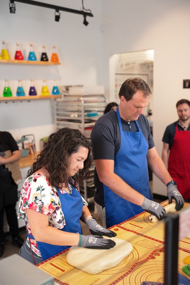 Two people in blue aprons wearing gloves stretch and roll warm taffy on a marked silicone mat in a bright candy-making workshop with colorful beakers on shelves in the background.