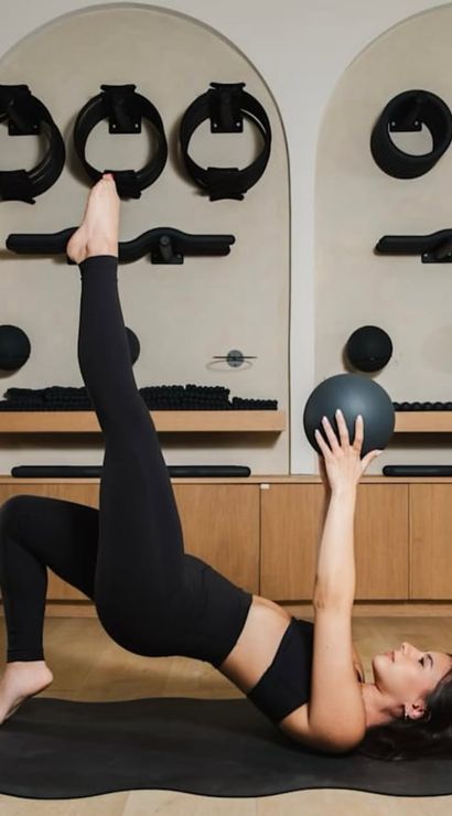Person doing a single-leg glute-bridge with one leg raised and holding a medicine ball overhead on a mat in a modern Pilates studio with wall-mounted fitness gear