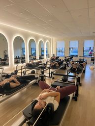 Group Pilates reformer class with participants reclining on reformer machines in a modern studio with arched backlit mirrors, light wood floors, and street-facing windows.