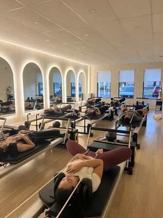 Group Pilates reformer class with participants reclining on reformer machines in a modern studio with arched backlit mirrors, light wood floors, and street-facing windows.