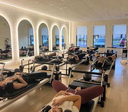 Group Pilates reformer class with participants reclining on reformer machines in a modern studio with arched backlit mirrors, light wood floors, and street-facing windows.