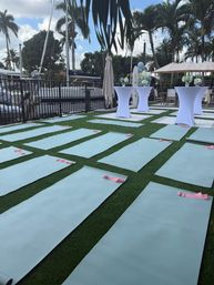 Outdoor waterfront yoga setup at a marina — rows of light-blue yoga mats with pink resistance bands on artificial grass, white cocktail tables, palm trees and sailboats in the background