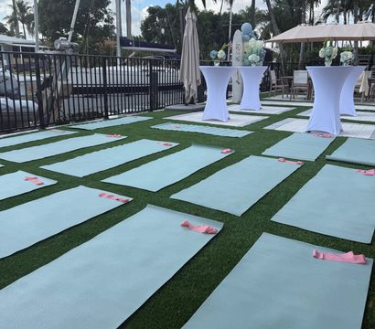 Outdoor waterfront yoga setup at a marina — rows of light-blue yoga mats with pink resistance bands on artificial grass, white cocktail tables, palm trees and sailboats in the background