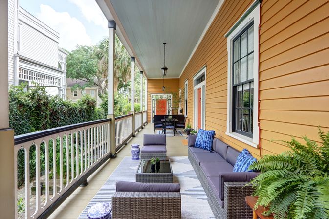 Cozy covered wraparound porch with warm orange siding, wicker sectional sofa with blue pillows, striped rug, coffee table, potted ferns and a dining area, overlooking a leafy residential yard.