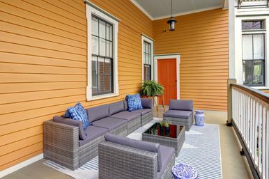 Cozy front porch with bright orange siding and red-orange door, gray wicker sectional and ottomans with slate cushions, navy patterned pillows, glass-top coffee table on a striped outdoor rug, potted fern and white railing.