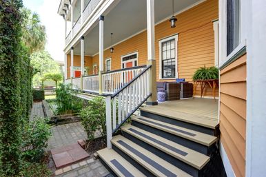 Cheerful orange clapboard house with an inviting covered porch—black-trimmed stairs and white railing, hanging lanterns, wicker seating, potted ferns, and a narrow garden water feature alongside a brick path.