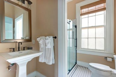 Bright, airy bathroom with pedestal sink and black matte faucet, framed mirror, white towels on a rail, glass walk-in shower with subway tile, frosted window with woven shade, and black-and-white hexagon floor tile.