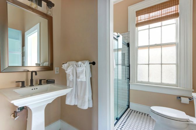 Bright, airy bathroom with pedestal sink and black matte faucet, framed mirror, white towels on a rail, glass walk-in shower with subway tile, frosted window with woven shade, and black-and-white hexagon floor tile.