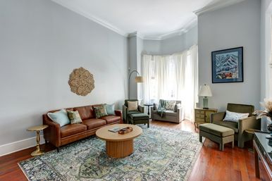 Sunlit bay-window living room with hardwood floors, brown leather sofa, patterned green area rug, round wooden coffee table, olive accent chairs and sheer white curtains
