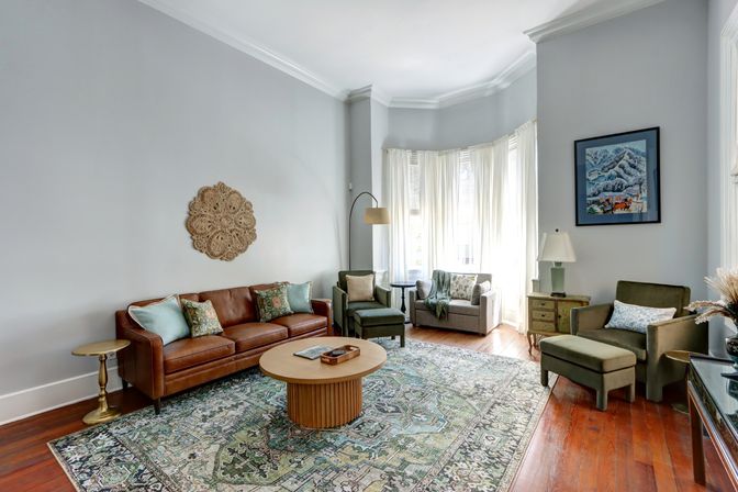 Sunlit bay-window living room with hardwood floors, brown leather sofa, patterned green area rug, round wooden coffee table, olive accent chairs and sheer white curtains