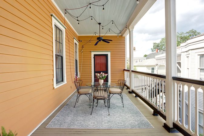 Charming covered second-story porch with warm orange siding, white railing, string lights and ceiling fan, round glass table and four wrought-iron chairs on a patterned outdoor rug